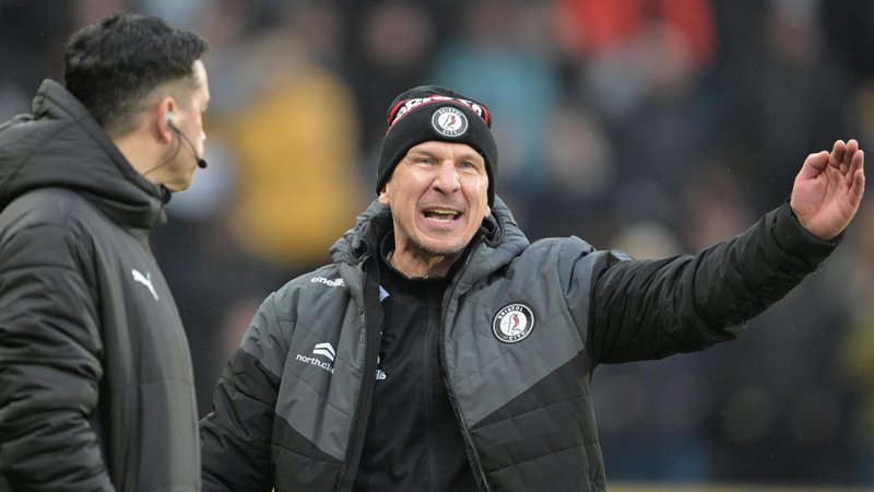 Bristol City head coach Gerhard Struber, wearing a black and grey jacket and black bobble hat, both with the Robins club badge on, angrily gesticulates towards the fourth official in a Championship game. The fourth official, wearing a black Puma jacket, looks at Struber and crowd behind Struber is blurred.