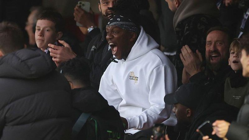 KSI celebrates after Dagenham & Redbridge score a goal, surrounded by supporters in the stand