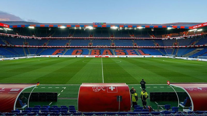 View of St Jakob-Park stadium taken from behind a red dugout on the halfway line, with the name FC Basel in red text on a blue background on the seats opposite