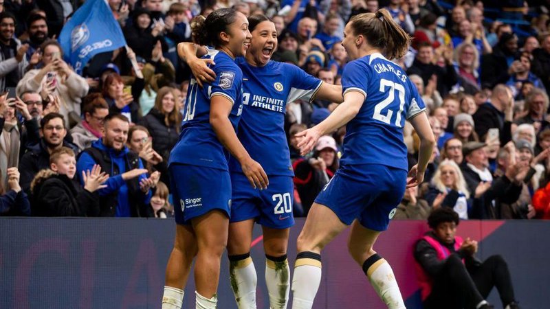 Lauren James celebrates scoring at Stamford Bridge as Sam Kerr and Niamh Charles join her in a huddle, with fans applauding in the stand behind them