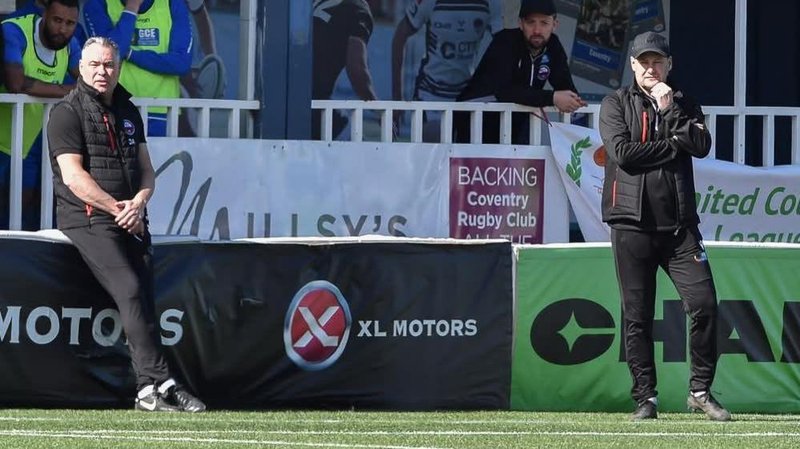 Darren Acton sits on an advertising board watching on with joint-manager Russell Dodd on the touchline during a match