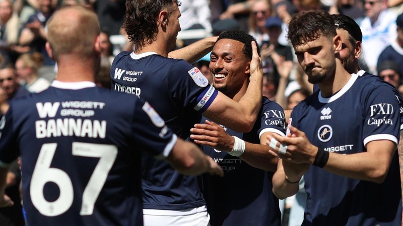 Femi Azeez, wearing the navy blue Millwall home shirt, smiles as he celebrates with his team-mates after scoring against Oxford. Jake Cooper is grabbing his head and the back of Barry Bannan can be seen in the foreground. Mihailo Ivanovic, stood to the right, is clapping his hands.