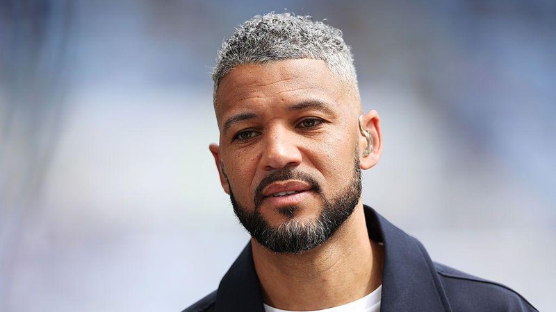 Jobi McAnuff, wearing a navy blue jacket, sporting a beard with tinges of grey, he also has short grey hair and is wearing a TV earpiece