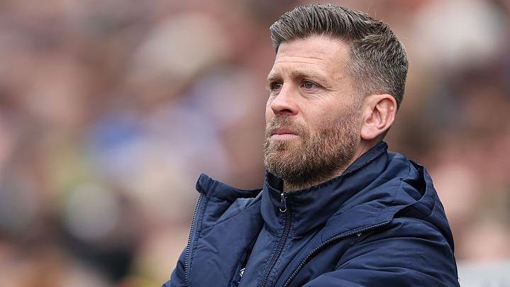 Boreham Wood manager Luke Garrard wearing a blue overcoat watching his side from the touchline at Carlisle