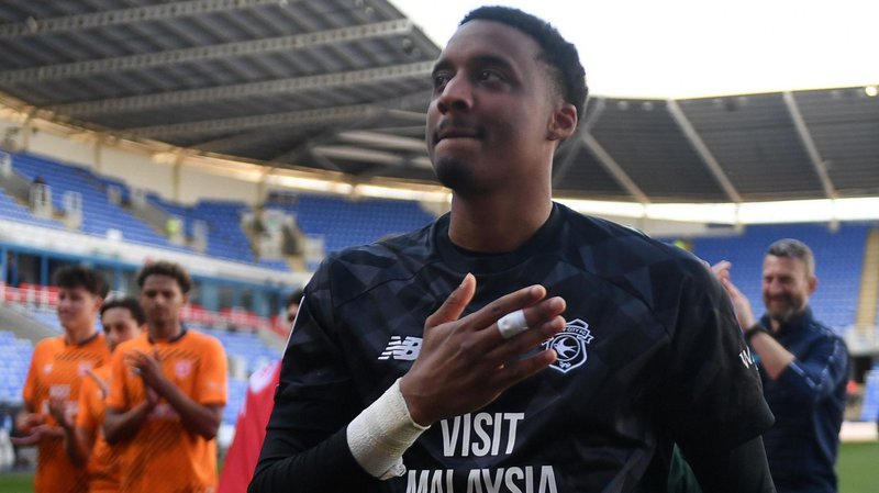 Nathan Trott in black Cardiff goalkeeper shirt, with his raised right hand partially over the club badge as figures including players behind him applaud