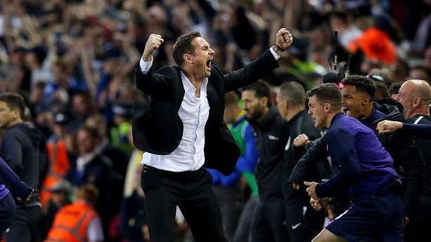 Frank Lampard celebrates on the touchline wearing a black suit and white shirt with fists clenches above his head