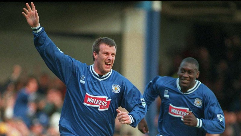 Steve Claridge celebrates scoring a goal for Leicester wearing a blue shirt with the club's badge of a fox on it. Claridge has his right arm raised in the air with teammate Emile Heskey chasing him.