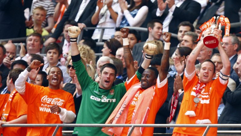 Charlie Adam of Blackpool in an orange shirt lifts the play-off trophy above his head while his team-mates raise their arms in celebration