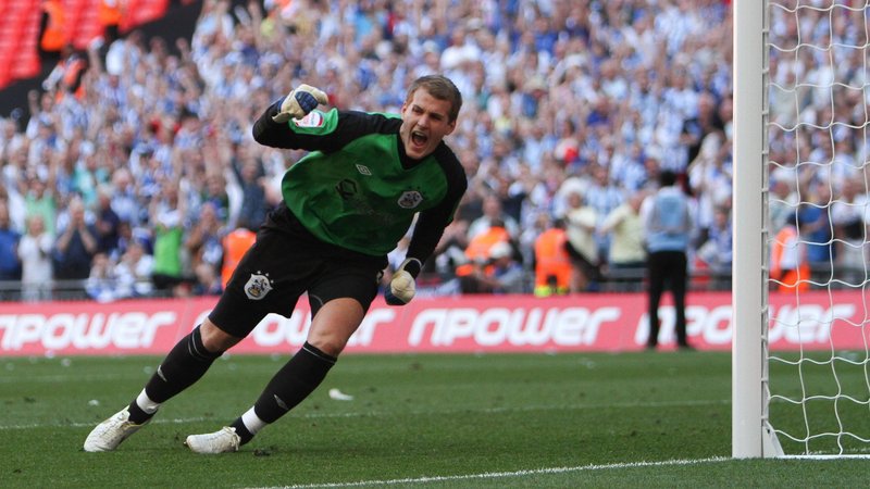 Huddersfield goalkeeper Alex Smithies wearing a green jersey clenches his right fist in celebrating saving a penalty