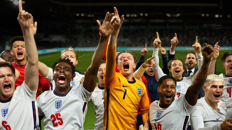 In a scene from Dear England, a team of footballers wearing the England men's team white and red kit celebrate on a football pitch, their goalkeeper, wearing yellow stands in the middle.