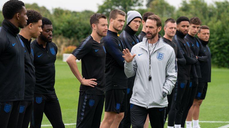 In a scene from Dear England, a man with brown hair wearing a silver windbreaker jacket stands in from of a team of football players on a feild wearing blue and black kit.