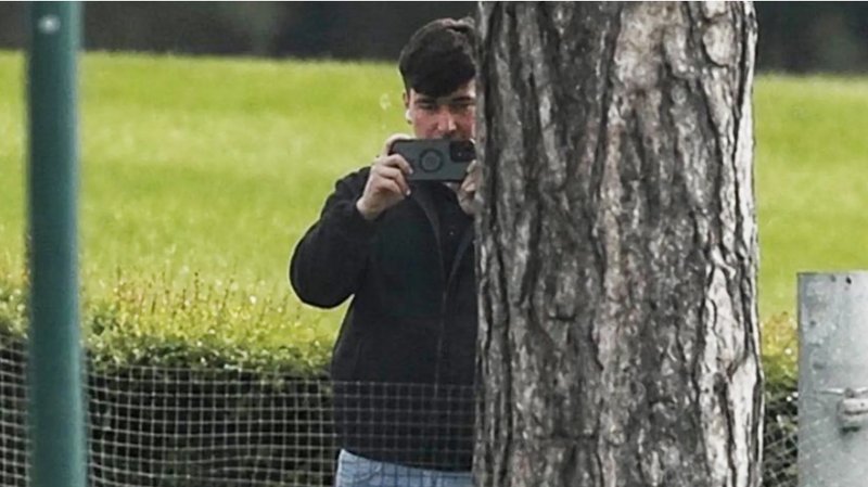 A man in a dark jacket and blue jeans stands partially concealed by a tree trunk and holds a phone up in two hands as if filming and has a white earphone in his right ear