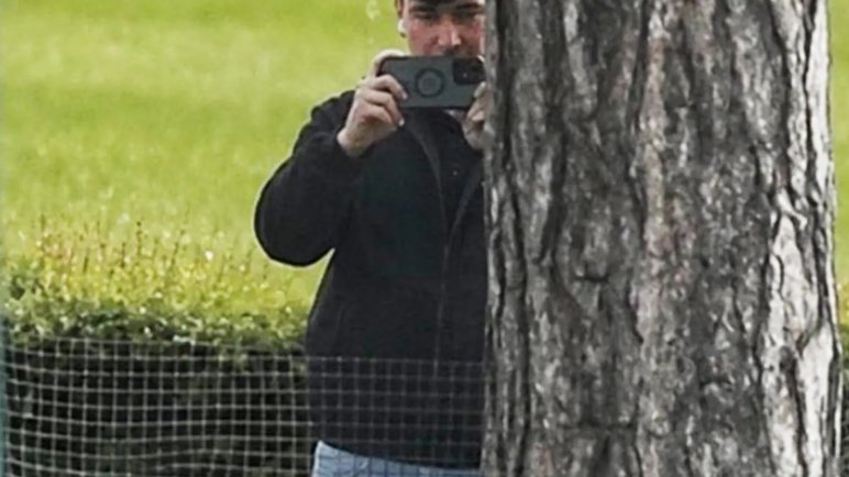 A man in a dark jacket and blue jeans stands partially concealed by a tree trunk and holds a phone up in two hands as if filming and has a white earphone in his right ear