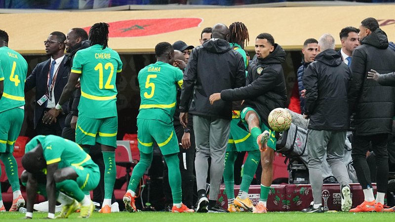 Senegal players and coaches stand on the touchline, many with their backs to the camera, during the 2025 Afcon final while protesting against a referee's decision, with Iliman Ndiaye facing the camera and seen midway through kicking a ball across the pitch in anger. Another Senegal player in the foreground is on all fours with his head hanging down disconsolately
