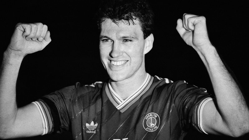 A black and white image of Charlton centre-back Peter Shirtliff punching his fists in the air either side of his head while smiling after the Addicks beat Leeds in the play-off final replay in May 1987. He can be seen wearing the Charlton home shirt with an Adidas logo and the Charlton club badge on the chest.