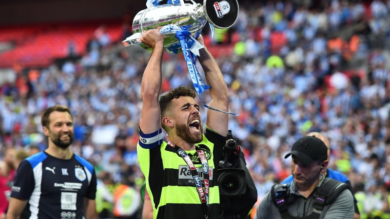 Tommy Smith, wearing a fluorescent green and black hooped Huddersfield away shirt, lifts the Championship play-off final trophy at Wembley in May 2017, Blue and white shirts of the Huddersfield fans in the stands can be seen behind him.